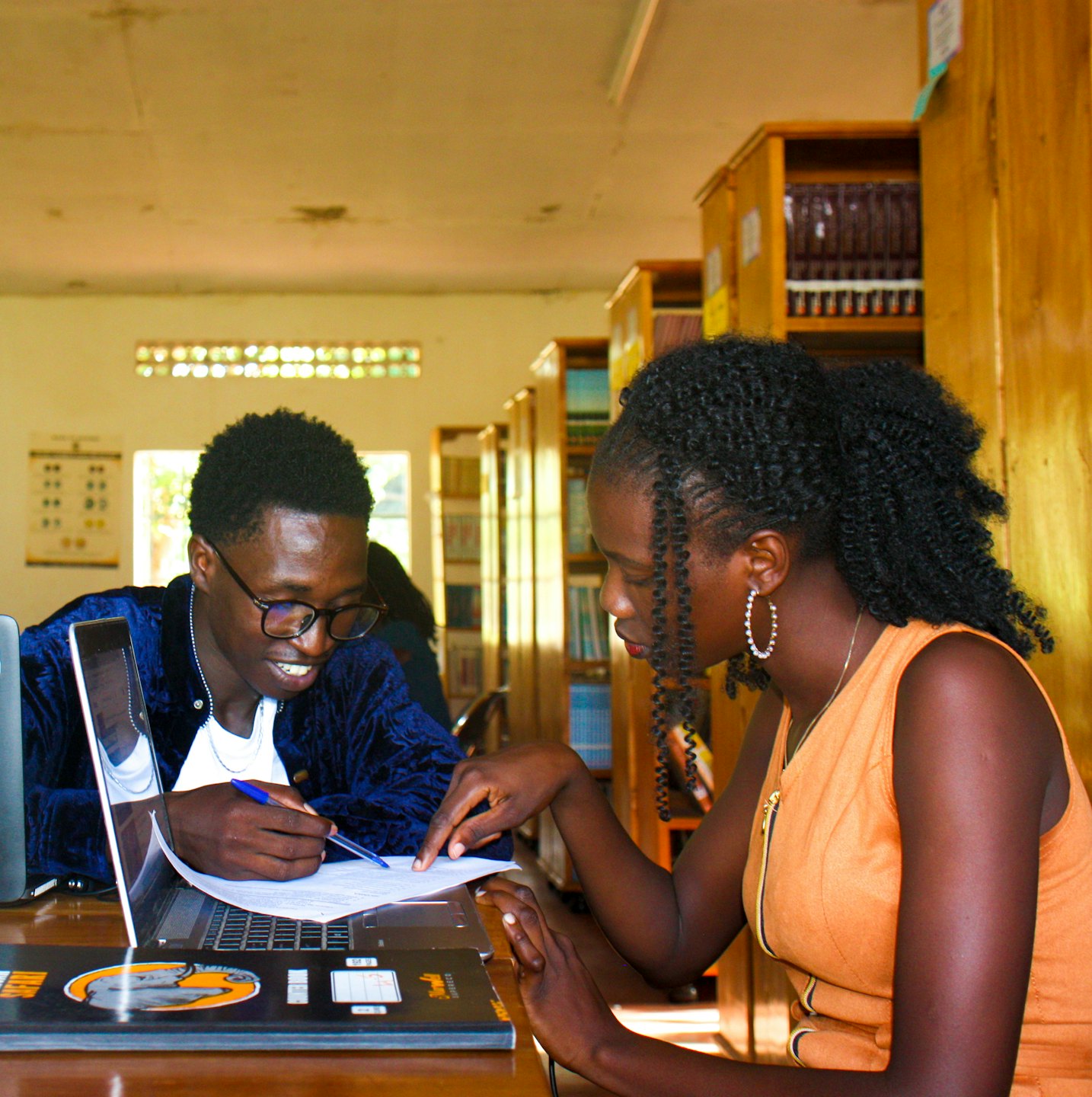 Two students discuss a document on a laptop in a library.