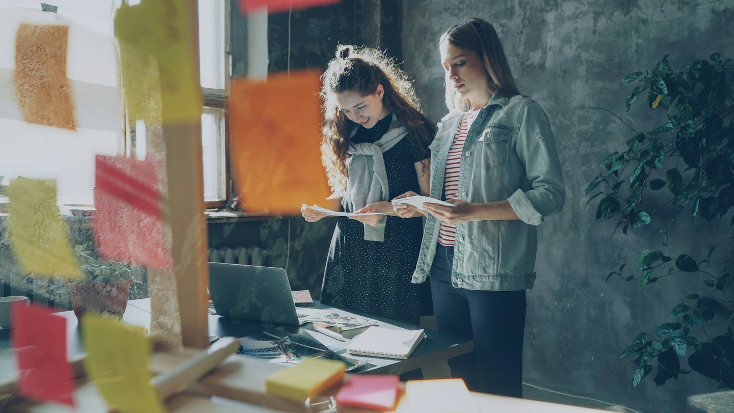Two women review documents and a laptop at an office desk.